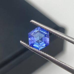 May include: A faceted, hexagonal blue gemstone is held by a pair of silver tweezers. The gem has a deep, saturated color and appears to be transparent. The background is a gradient of black and white.