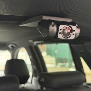 Puede incluir: Una cámara de salpicadero negra montada en el techo interior de un vehículo mediante un soporte metálico. La cámara tiene una lente y pequeñas luces. El interior del coche es visible.