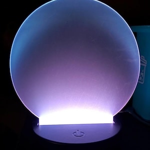 May include: A glowing, circular LED night light with a power button. The light emits a gradient of purple and blue hues, with a bright white glow at the base. The base is black and has a power symbol.