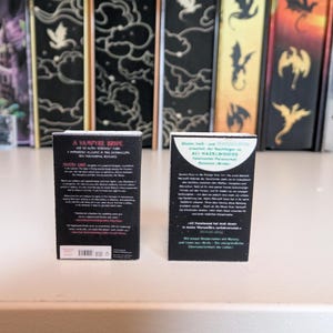 May include: Two miniature book replicas standing upright. The left book has a black cover with white text titled A VAMPYRE BRIDE. The right book has a black cover with a white circular graphic and German text. Both items feature detailed back cover text.