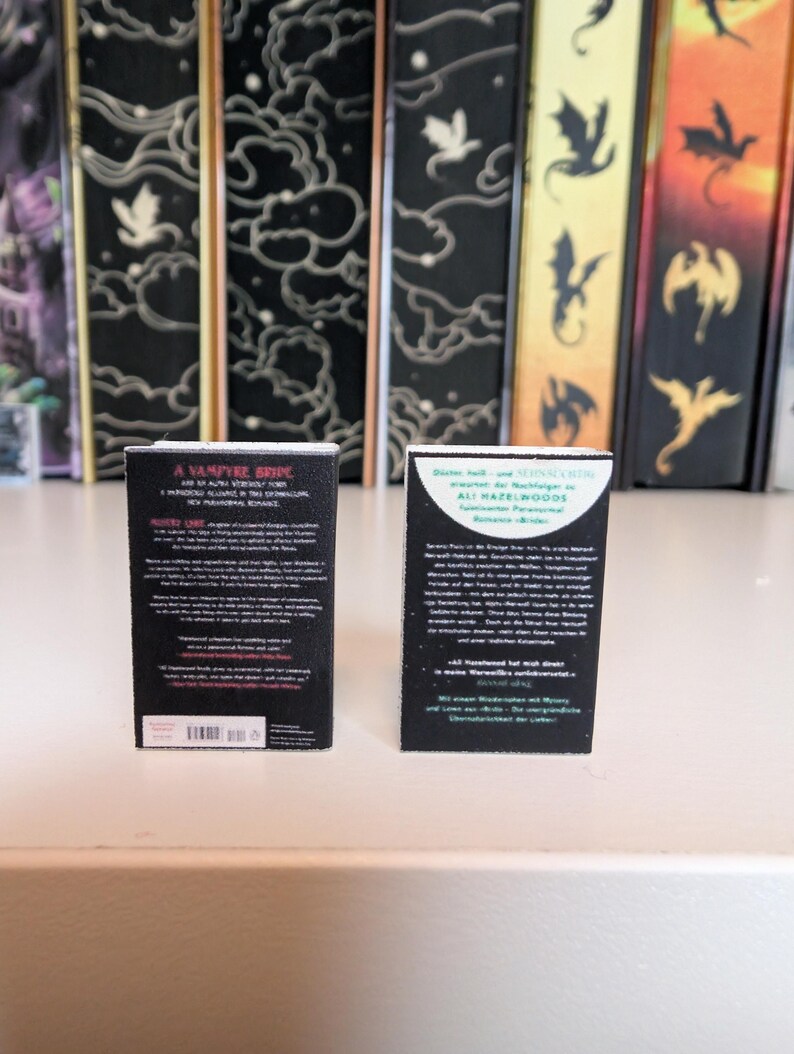 May include: Two miniature book replicas standing upright. The left book has a black cover with white text titled A VAMPYRE BRIDE. The right book has a black cover with a white circular graphic and German text. Both items feature detailed back cover text.