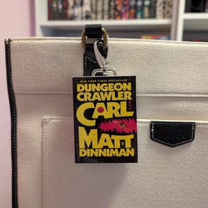 May include: A rectangular bag charm featuring the book cover art for Dungeon Crawler Carl by Matt Dinniman, with yellow text on a black background, attached to a silver-toned metal lobster clasp hanging from a cream-coloured canvas tote bag.
