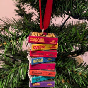 May include: A miniature stack of Dungeon Crawler Carl books by Matt Dinniman, featuring titles like The Gate of the Feral Gods and The Butcher's Masquerade, suspended by a red ribbon. The colourful book spines are stacked vertically as a hanging ornament.