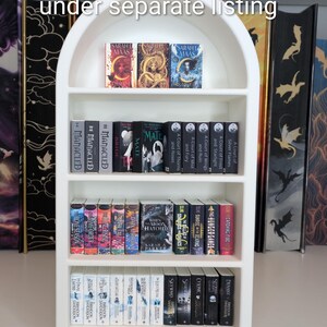 May include: A white arched miniature bookshelf filled with rows of tiny book replicas. The text "Mini bookshelf available under separate listing" is printed at the top. The shelves contain various miniature book spines with titles and cover art.