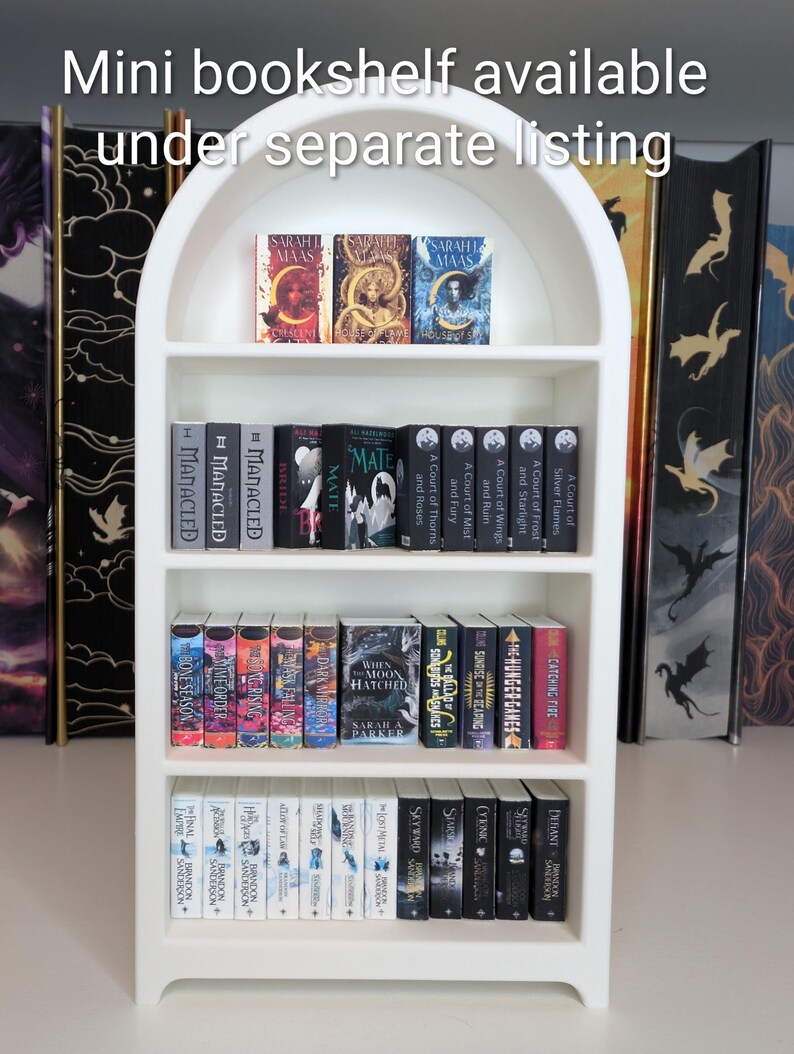 May include: A white arched miniature bookshelf filled with rows of tiny book replicas. The text "Mini bookshelf available under separate listing" is printed at the top. The shelves contain various miniature book spines with titles and cover art.