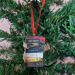 May include: A miniature stack of six books hanging from a red ribbon, featuring the titles Red Rising, Golden Son, Morning Star, Iron Gold, Dark Age, and Light Bringer. Each book spine displays its respective title and author name in clear text.