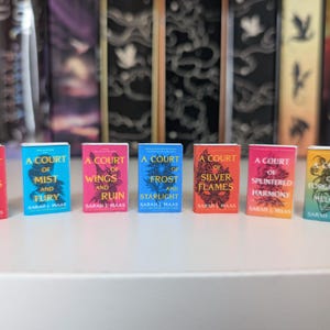 May include: Seven miniature books by Sarah J. Maas arranged in a row. Titles include A Court of Thorns and Roses, A Court of Mist and Fury, A Court of Wings and Ruin, A Court of Frost and Starlight, A Court of Silver Flames, A Court of Splintered Harmony, and A Court of Forgotten Melody.