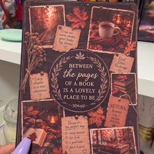 May include: A rectangular decorative print featuring autumn-themed imagery and text. The central text reads: BETWEEN the pages OF A BOOK IS A LOVELY PLACE TO BE. Surrounding images include fallen leaves, books, a mug, and a lantern with notes.