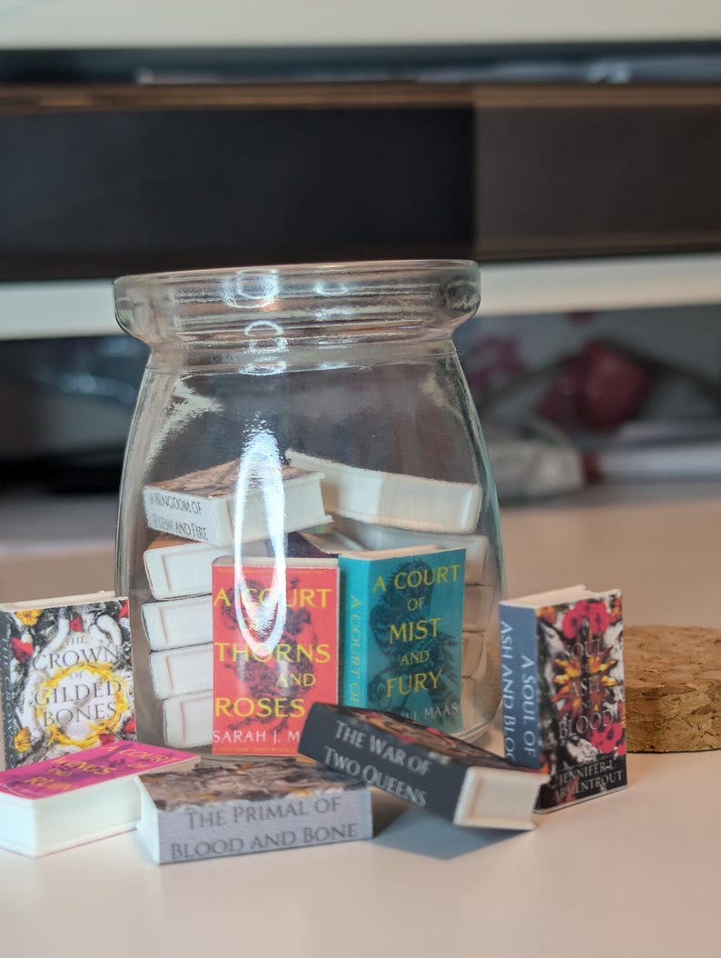 May include: A clear glass jar filled with miniature book replicas, including titles like A Court of Thorns and Roses, A Court of Mist and Fury, The Crown of Gilded Bones, The War of Two Queens, and A Soul of Ash and Blood, with a cork lid nearby.