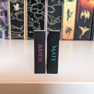 May include: Two rectangular black blocks standing upright. The left block features the word BRIDE in pink lettering, while the right block features the word MATE in green lettering. The blocks are positioned in front of several vertical book spines.