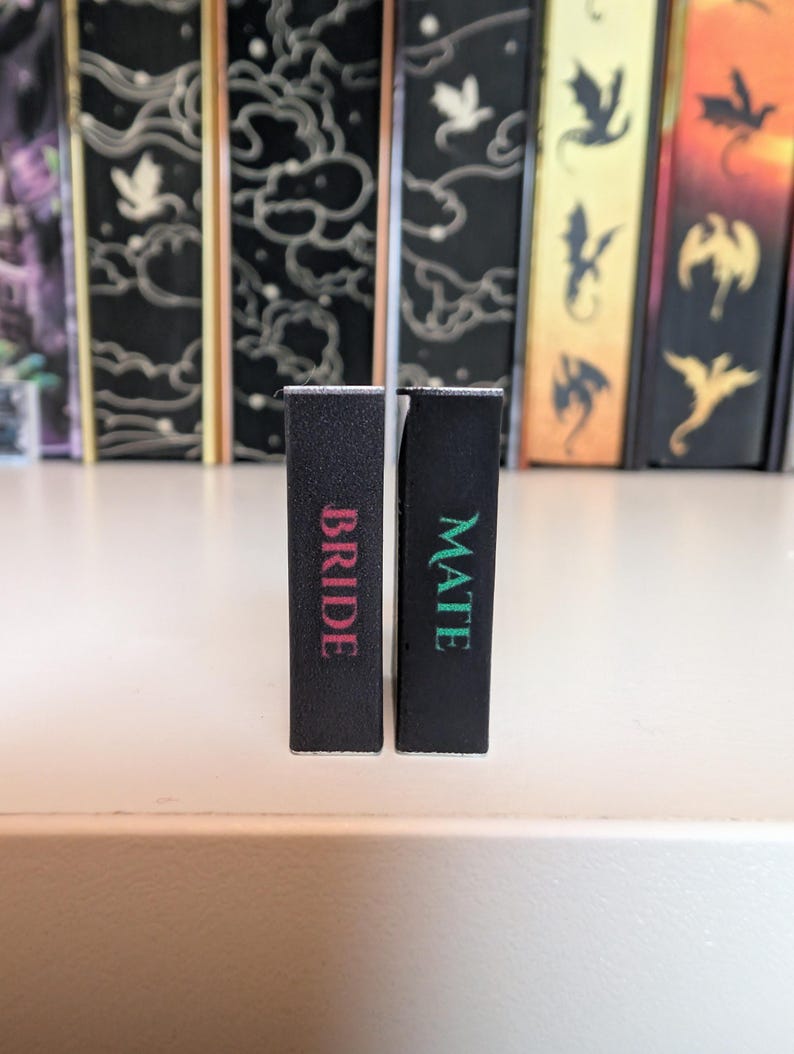 May include: Two rectangular black blocks standing upright. The left block features the word BRIDE in pink lettering, while the right block features the word MATE in green lettering. The blocks are positioned in front of several vertical book spines.