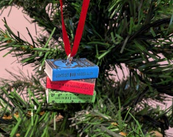 May include: A miniature ornament featuring a stack of three books suspended by a red ribbon. The top book is blue with the text "LIGHTS OUT" and "NAVESSA ALLEN". The middle book is red with "CAUGHT UP" and "NAVESSA ALLEN". The bottom book is green with "GAME ON" and "NAVESSA ALLEN".