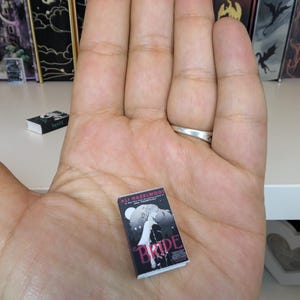 May include: A miniature book titled Bride by Ali Hazelwood with a black cover featuring a white illustration of a figure and a full moon. The book is held in an open palm, showcasing its small scale relative to the hand, with white pages visible.