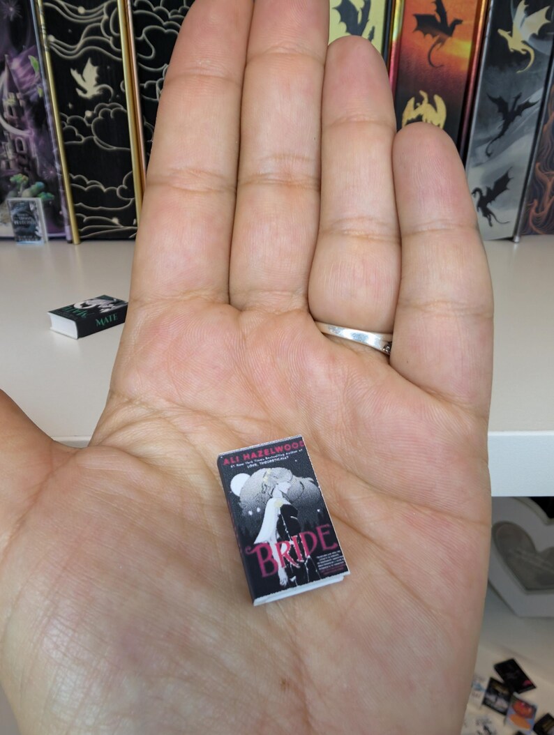 May include: A miniature book titled Bride by Ali Hazelwood with a black cover featuring a white illustration of a figure and a full moon. The book is held in an open palm, showcasing its small scale relative to the hand, with white pages visible.