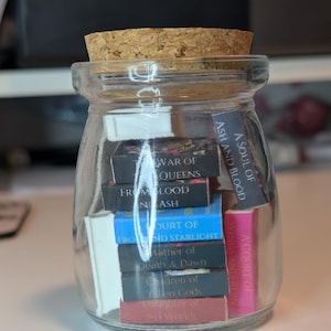 May include: A glass jar with a cork stopper, containing small books with colourful spines. Titles include "War of Queens" and "A Court of Wings and Ruin". The jar is on a light surface. The jar is clear.