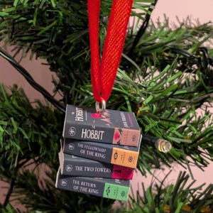 May include: A miniature book ornament featuring a stack of four J.R.R. Tolkien titles: The Hobbit, The Fellowship of the Ring, The Two Towers, and The Return of the King. The stack is suspended by a bright red ribbon attached to a silver metal loop.
