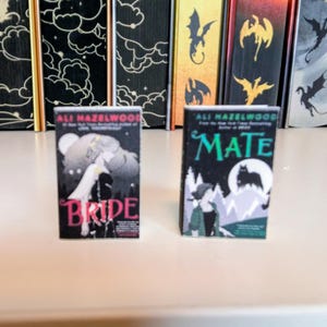 May include: Two miniature book replicas featuring the titles Bride and Mate by author Ali Hazelwood. The Bride cover is black with pink text and a wolf illustration, while the Mate cover is black and green with white text and a wolf silhouette.