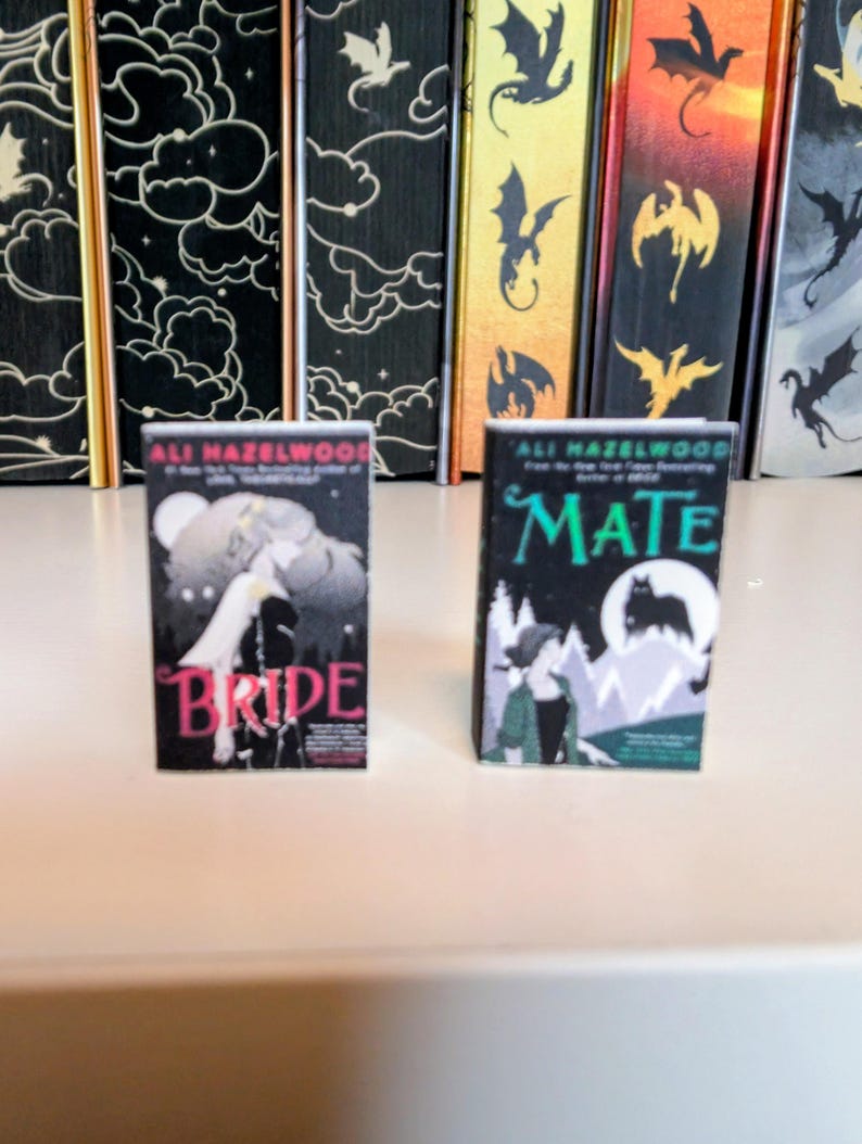 May include: Two miniature book replicas featuring the titles Bride and Mate by author Ali Hazelwood. The Bride cover is black with pink text and a wolf illustration, while the Mate cover is black and green with white text and a wolf silhouette.