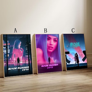 May include: Three framed Blade Runner 2049 film posters. Each poster displays a different image from the movie, with vivid colours and the film's title clearly visible. The posters are in wooden frames and are leaning against a wall.