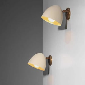 Puede incluir: Dos apliques de pared color crema con acabado de latón. Los apliques tienen forma curva de lágrima con interior amarillo cálido. Están montados en una pared blanca, con una pared gris en el fondo.