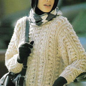 Könnte beinhalten: Ein cremefarbener Zopfstrickpullover mit Kapuze und einem schwarz-weiß karierten Schal. Die Person trägt schwarze Handschuhe und eine schwarze Umhängetasche mit Karomuster.