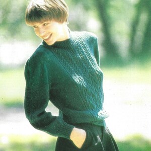 May include: A dark green knitted jumper with a textured cable knit pattern on the front. The jumper has long sleeves and a crew neck. The person is wearing black trousers and has short brown hair.