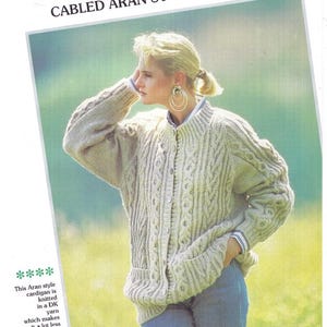 Pu&ograve; includere: Un cardigan beige in stile Aran a maglia a treccia con chiusura a bottoni. Il maglione presenta intricati motivi a treccia e viene indossato con jeans blu. L'immagine include il testo "CABLED ARAN STYLE CARDIGAN".