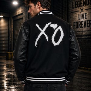 May include: A black varsity jacket featuring long black leather sleeves and a white embroidered XO logo with a heart on the back. The jacket includes a ribbed collar and cuffs with white stripes, and it is worn over dark denim jeans.