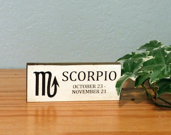 May include: A rectangular wooden sign with the word "SCORPIO" in black letters, along with the dates October 23 - November 21. The sign also features the Scorpio zodiac symbol. The sign is on a wooden surface.