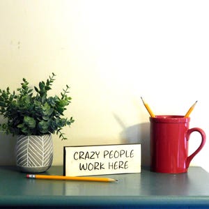 May include: A wooden sign with the text "CRAZY PEOPLE WORK HERE" in black marker. The sign is sitting on a teal surface next to a red mug with two yellow pencils sticking out of it. A small potted plant with green leaves is in the background.
