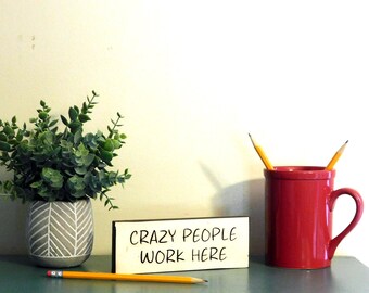 May include: A wooden sign with the text "CRAZY PEOPLE WORK HERE" in black marker. The sign is sitting on a teal surface next to a red mug with two yellow pencils sticking out of it. A small potted plant with green leaves is in the background.