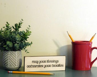 May include: A decorative arrangement featuring a small potted plant, a wooden sign with the text "may your blessings outnumber your troubles," a red mug, and pencils. The items are arranged on a teal surface against a cream-colored wall.