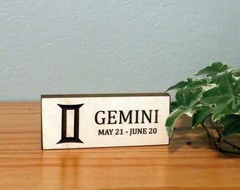 May include: A wooden block sign with the word "GEMINI" in black text, along with the dates "MAY 21 - JUNE 20". The sign features a black Gemini symbol. The sign is on a wooden surface with a green plant in the background.