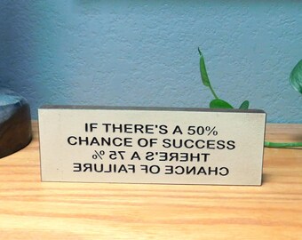 May include: A rectangular wooden block with the text "IF THERE'S A 50% CHANCE OF SUCCESS" printed in black. Below, the same text is printed in reverse. The block is beige and sits on a wooden surface.
