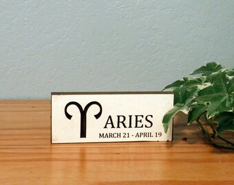 May include: A rectangular wooden sign with the word "ARIES" in black, along with the Aries symbol and the dates "MARCH 21 - APRIL 19." The sign is set on a wooden surface, with green and white leafy plants in the background.