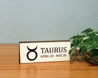 May include: A wooden sign with the Taurus zodiac symbol and text. The sign is rectangular with a light-colored front and dark edges. The text reads "TAURUS APRIL 20 - MAY 20" in black. The sign is on a wooden surface.