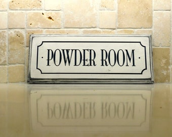 May include: A white wooden sign with black lettering that reads "POWDER ROOM" on a beige tiled wall. The sign is reflected in the surface below.