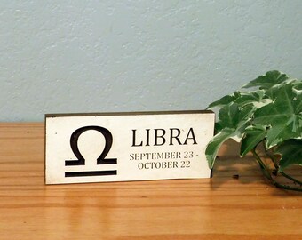 May include: A wooden sign with the Libra zodiac symbol and text. The sign displays the Libra symbol, the word "LIBRA", and the dates "SEPTEMBER 23 - OCTOBER 22". The sign is on a wooden surface with a plant in the background.