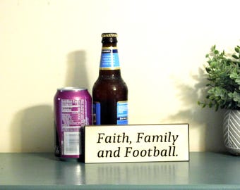 May include: A wooden sign with the text "Faith, Family and Football." The sign is sitting on a table with a can of soda and a bottle of beer.
