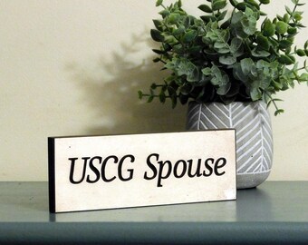 May include: White wooden sign with black text that reads "USCG Spouse". The sign is sitting on a blue surface in front of a potted plant.