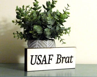 May include: A white wooden sign with black text that reads "USAF Brat". The sign is sitting on a green surface in front of a potted plant.