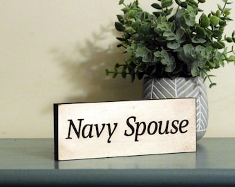 May include: A white wooden sign with black lettering that reads "Navy Spouse". The sign is sitting on a light blue surface.