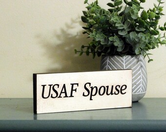 May include: A rectangular wooden sign with the text "USAF Spouse" in black lettering. The sign is off-white with a black border. A decorative plant in a gray and white patterned pot is in the background.