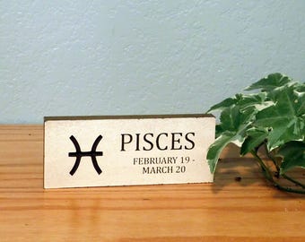May include: A wooden block sign with the word "PISCES" in black text, along with the Pisces symbol and the dates February 19 - March 20. The sign is on a wooden surface with green and white leafy plants in the background.