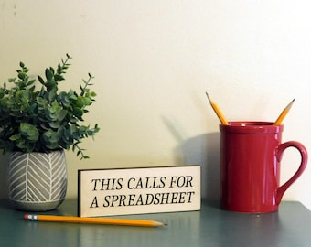 May include: A wooden sign with the text "This calls for a spreadsheet" in black lettering on a light brown background. The sign is sitting on a blue surface with a red mug and a potted plant.