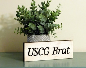 May include: A white wooden sign with black text that reads "USCG Brat". The sign is sitting on a teal surface with a potted plant in the background.