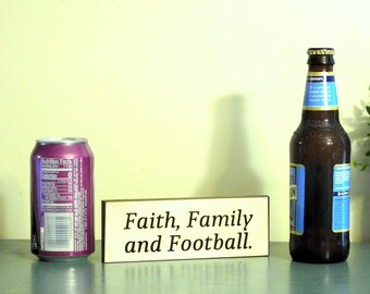 May include: A wooden sign with the text "Faith, Family and Football." The sign is sitting on a table with a can of soda and a bottle of beer.