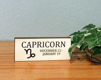 May include: A rectangular wooden sign with the word "CAPRICORN" in black letters, along with the Capricorn symbol and the dates December 22 - January 19. The sign is on a wooden surface with green and white leafy plants in the background.