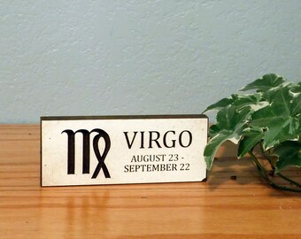 May include: A rectangular wooden sign with the Virgo astrological symbol and text. The sign is cream-colored with black lettering and a dark brown border. The text reads "VIRGO AUGUST 23 - SEPTEMBER 22". A green and white plant is in the background.