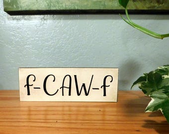 May include: A rectangular wooden sign with the text "f-CAW-f" in black cursive lettering. The sign is light-colored and sits on a wooden surface. Green plants are visible in the background.